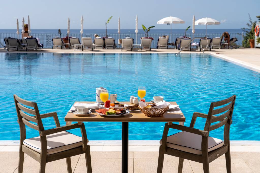 157 Mesa de café da manhã à beira da piscina do hotel Reid's Palace, na Ilha da Madeira, com duas cadeiras e espreguiçadeiras ao longe