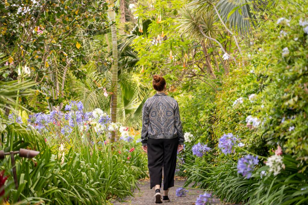 163 Mulher caminha pelo jardim do hotel Reid's Palace, na Ilha da madeira, entre plantas verdes, flores roxas e árvores