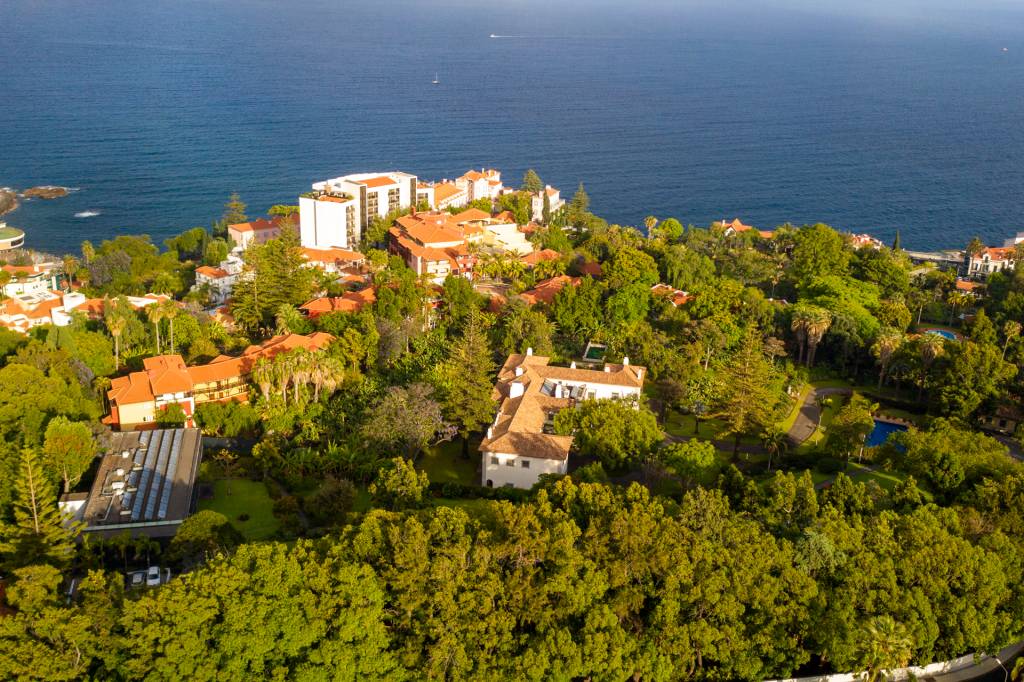 Imagem aérea de uma grande mancha verde de vegetação e um casarão branco no meio, com o mar e prédios ao fundo