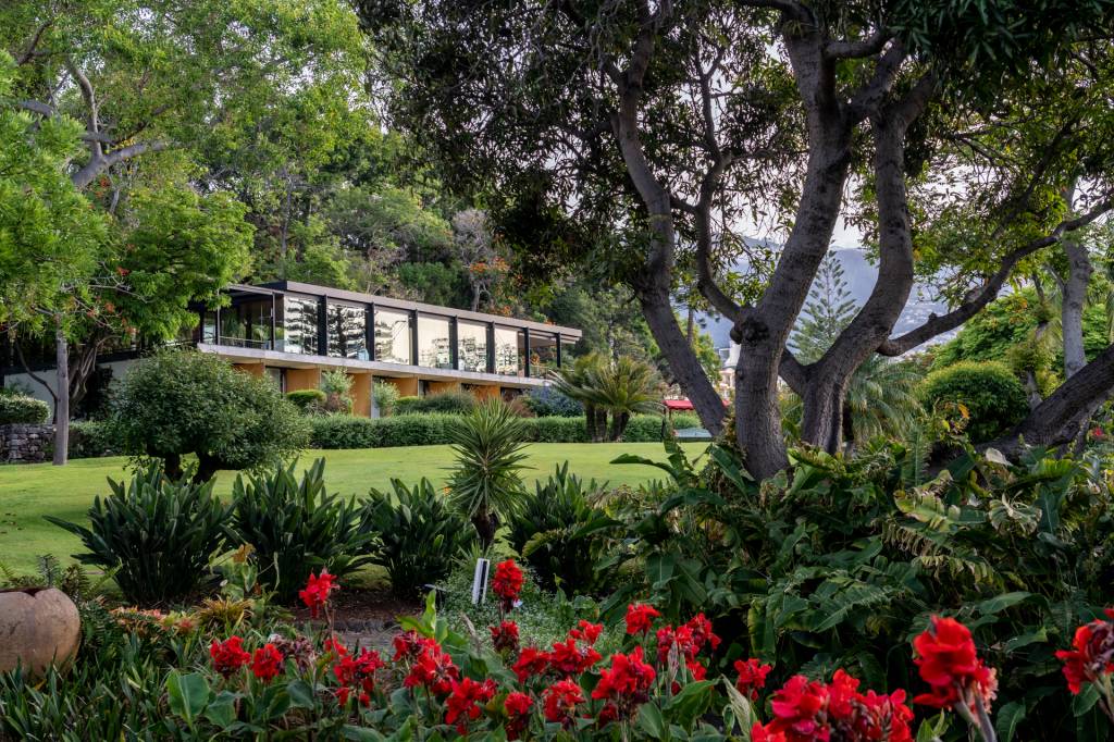 Flores vermelhas, arbustos e árvores com um gramado e uma construção de vidro ao fundo