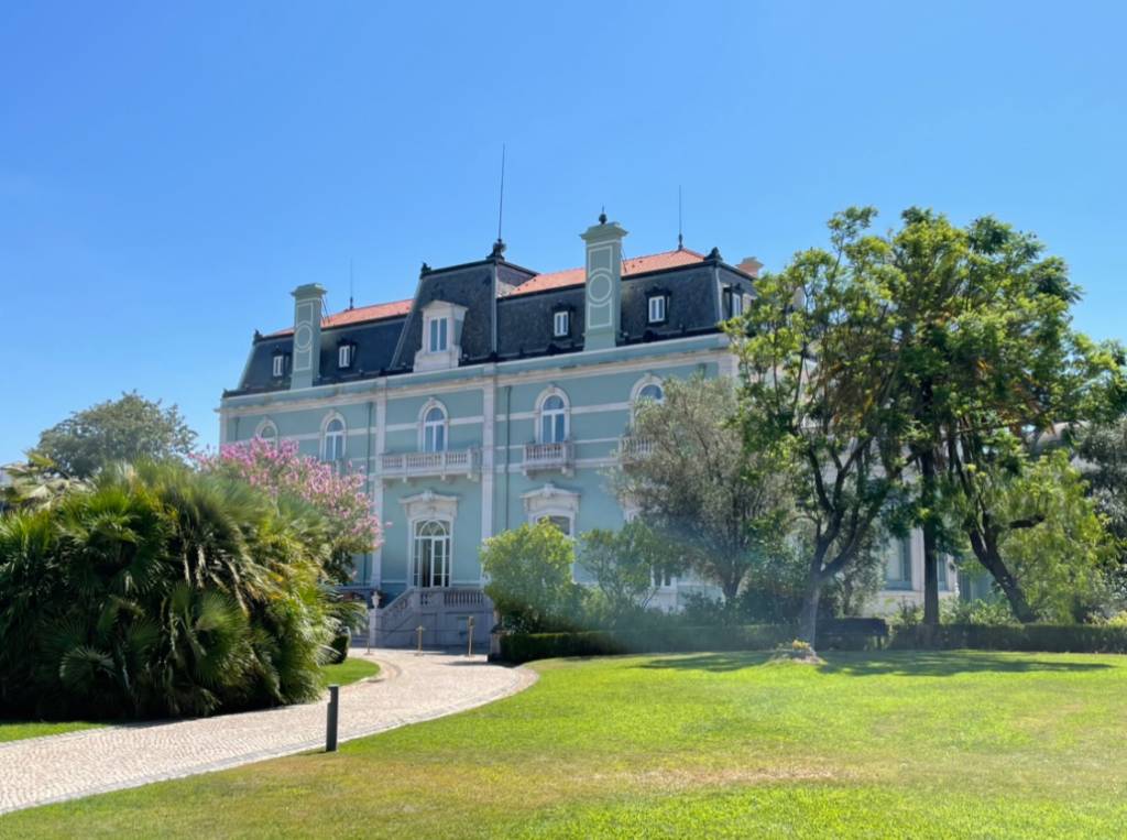 Gramado com árvores e, ao fundo, um palacete verde-água de três andares, com telhado cinzento