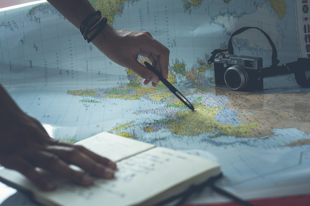 Pessoa usando caneta para apontar um local no mapa mundi enquanto segura um caderno de anotações
