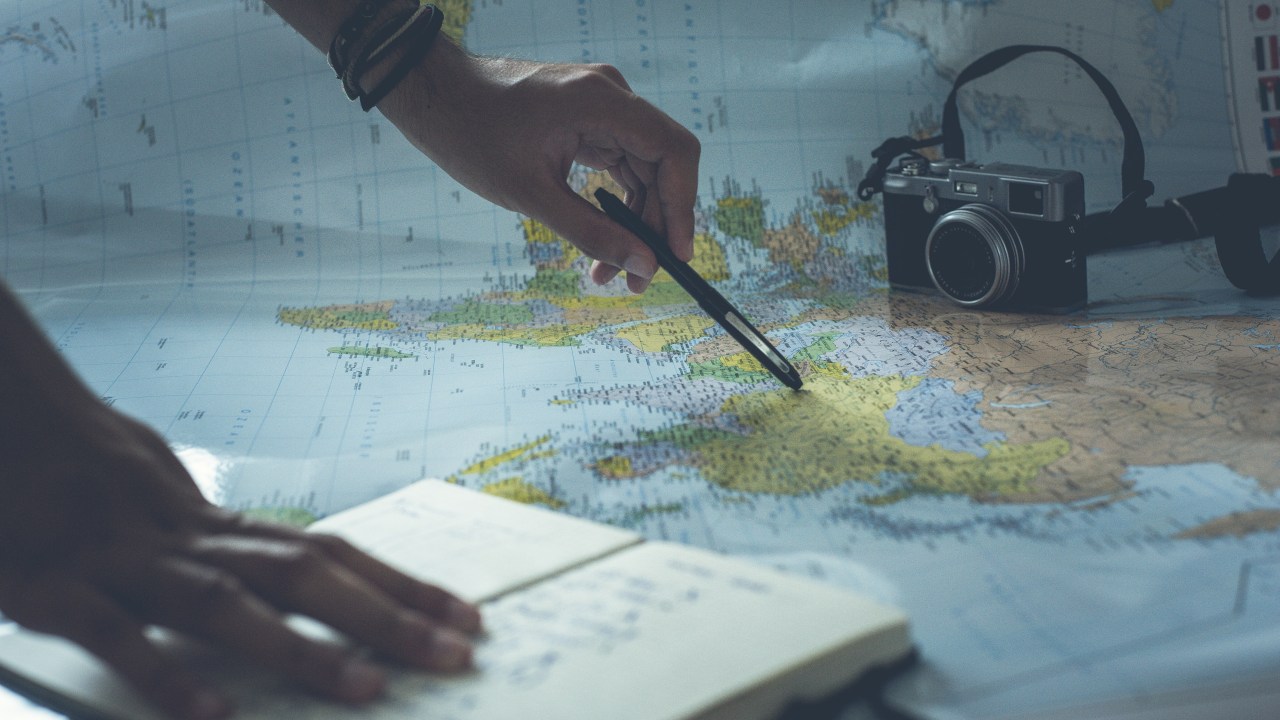 Pessoa usando caneta para apontar um local no mapa mundi enquanto segura um caderno de anotações