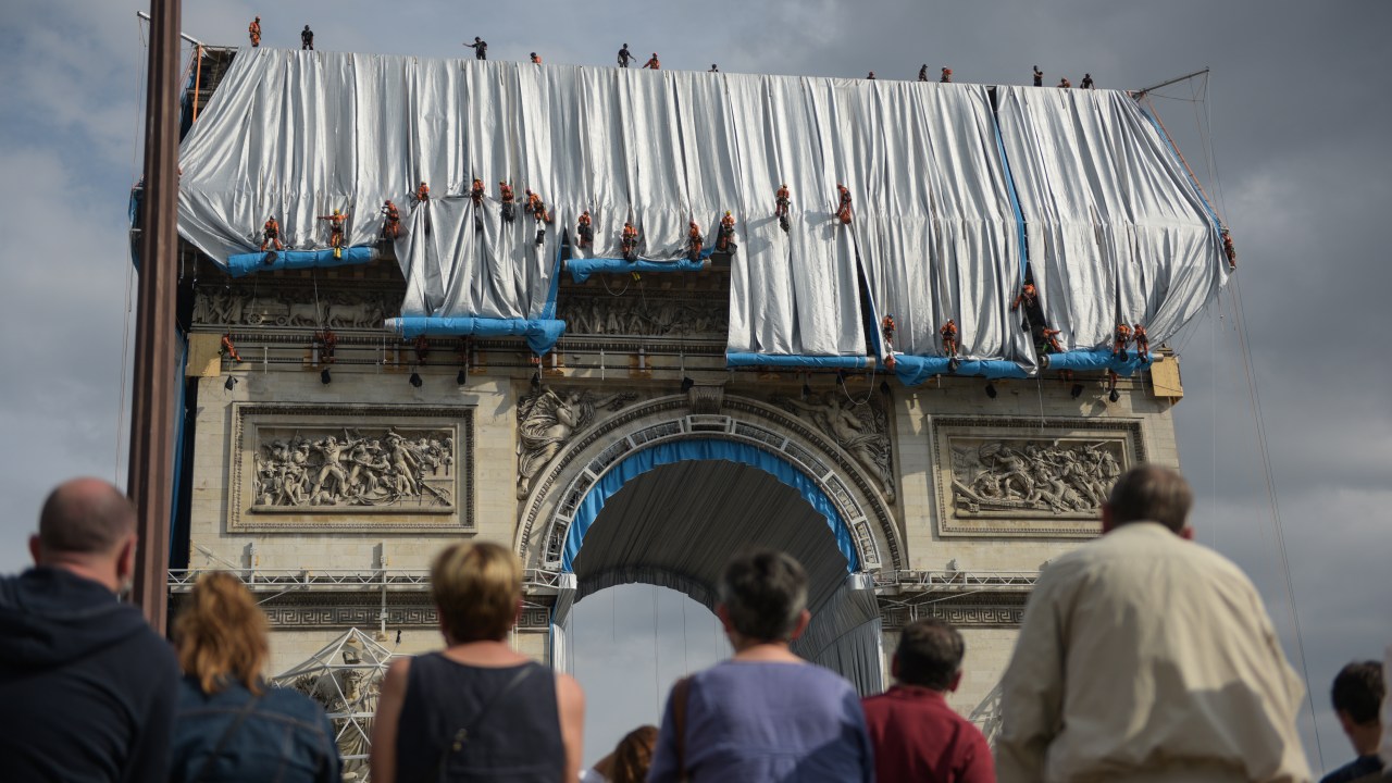 A imagem é uma foto tirada de baixo para cima. Ela mostra alguns trabalhadores colocando um pano sobre o Arco do Triunfo, em Paris. É possível ver que uma multidão parou para observar a obra.