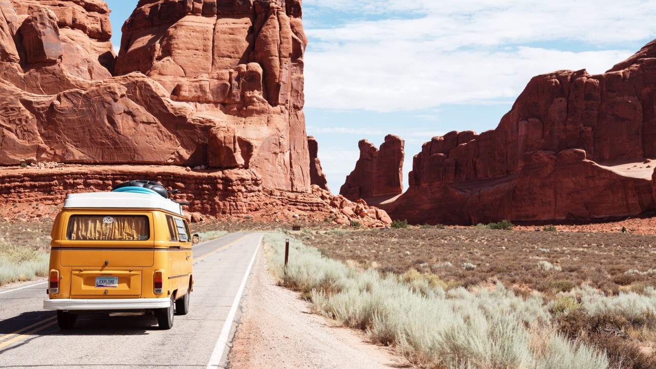 A imagem é uma foto de uma estrada no deserto, com uma kombi vista de costas, como se estivesse se encaminhando para as montanhas no fundo.