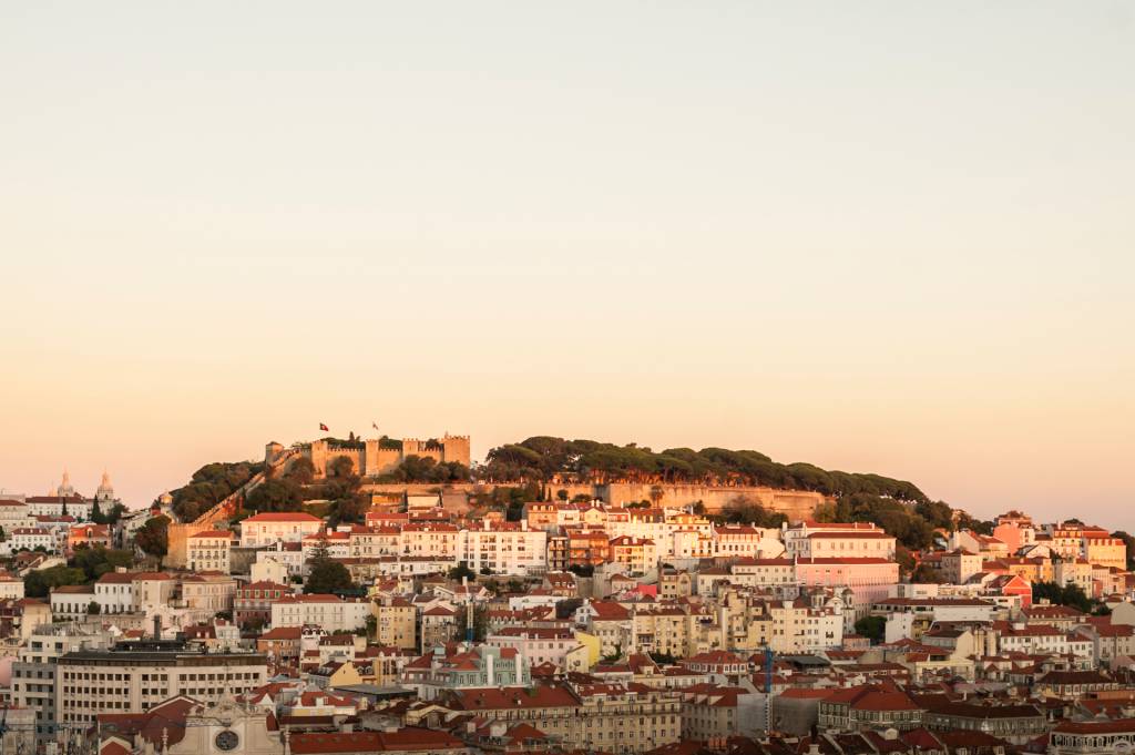 O casario de Lisboa a subir uma colina e, no alto, um castelo cercado de muralhas, ao pôr do sol