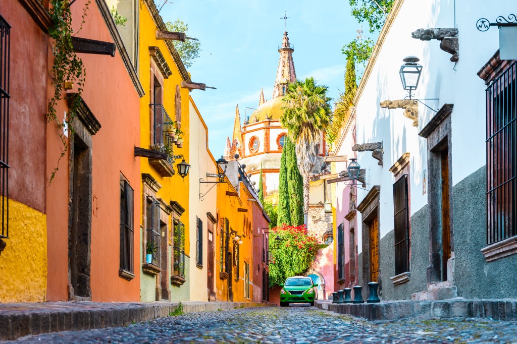A imagem mostra uma rua estreita em San Miguel de Allende, no Méxicoo. É possível ver construções colocaridas características em amarelo, rosa e branco. Ao fundo há uma construção que se assemelha /á uma catedral.