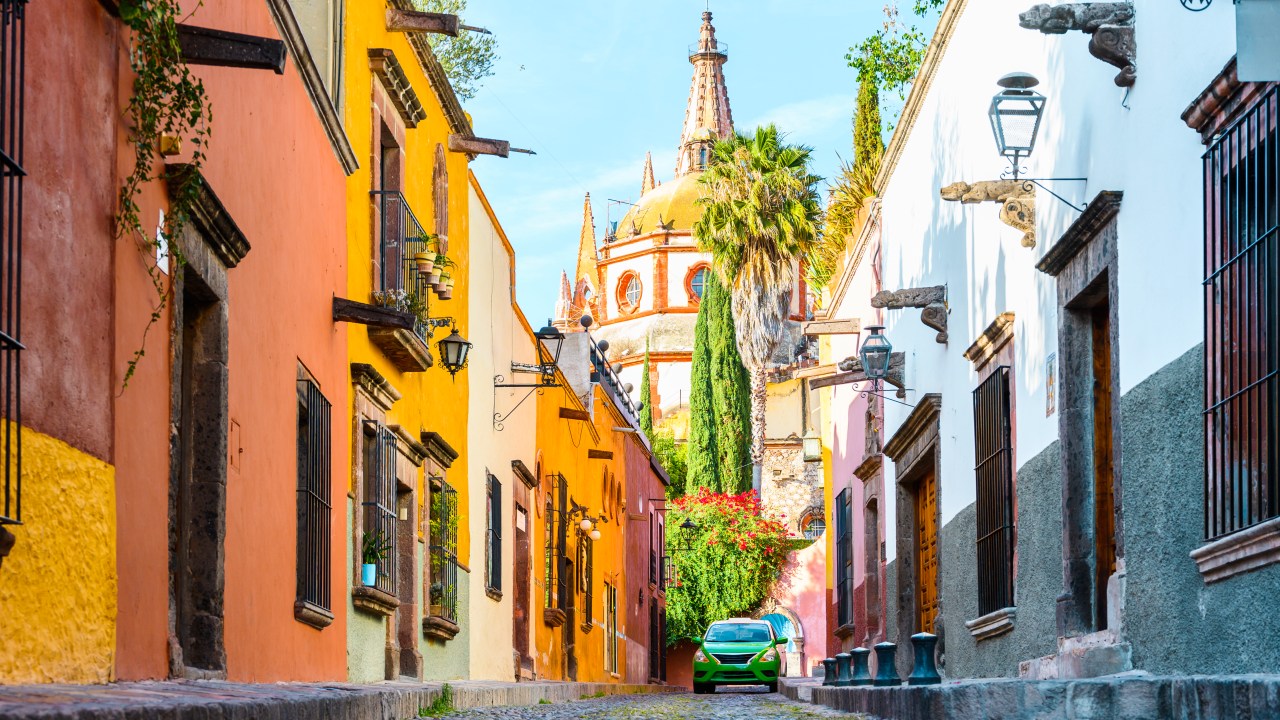 A imagem mostra uma rua estreita em San Miguel de Allende, no Méxicoo. É possível ver construções colocaridas características em amarelo, rosa e branco. Ao fundo há uma construção que se assemelha /á uma catedral.