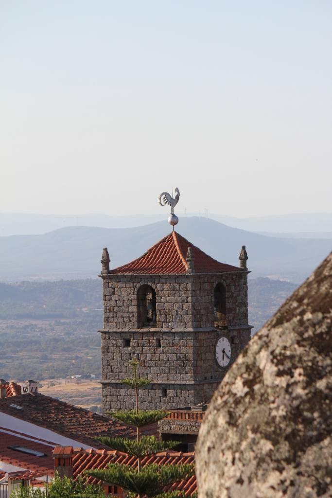 Uma torre de pedra com um galo no telhado, uma grande pedra desfocada em primeiro plano e colinas ao fundo