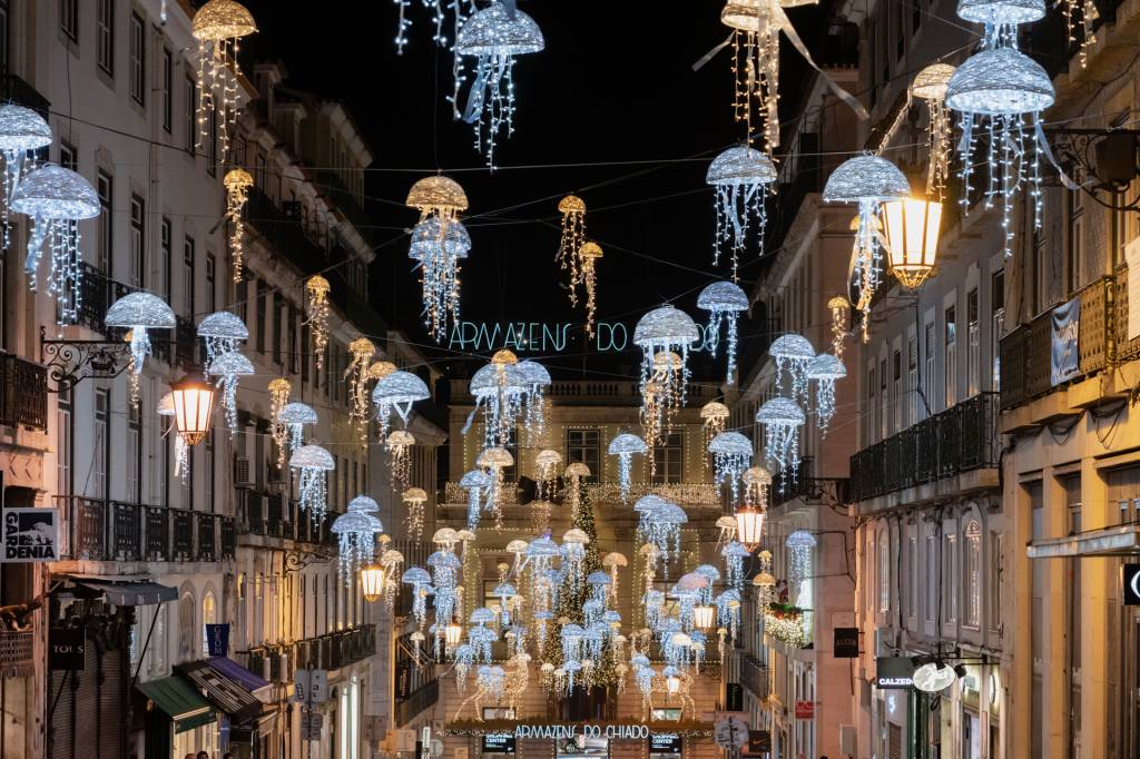Rua estreita, à noite, com dezenas de medusas de luz em azul e branco como enfeites de Natal