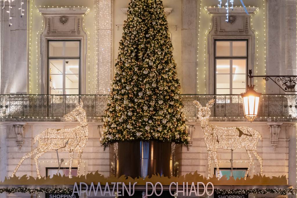 Grande árvore de Natal verde com decoração em branco e dourado e duas renas de luz ao lado, na fachada de um prédio, entre duas janelas