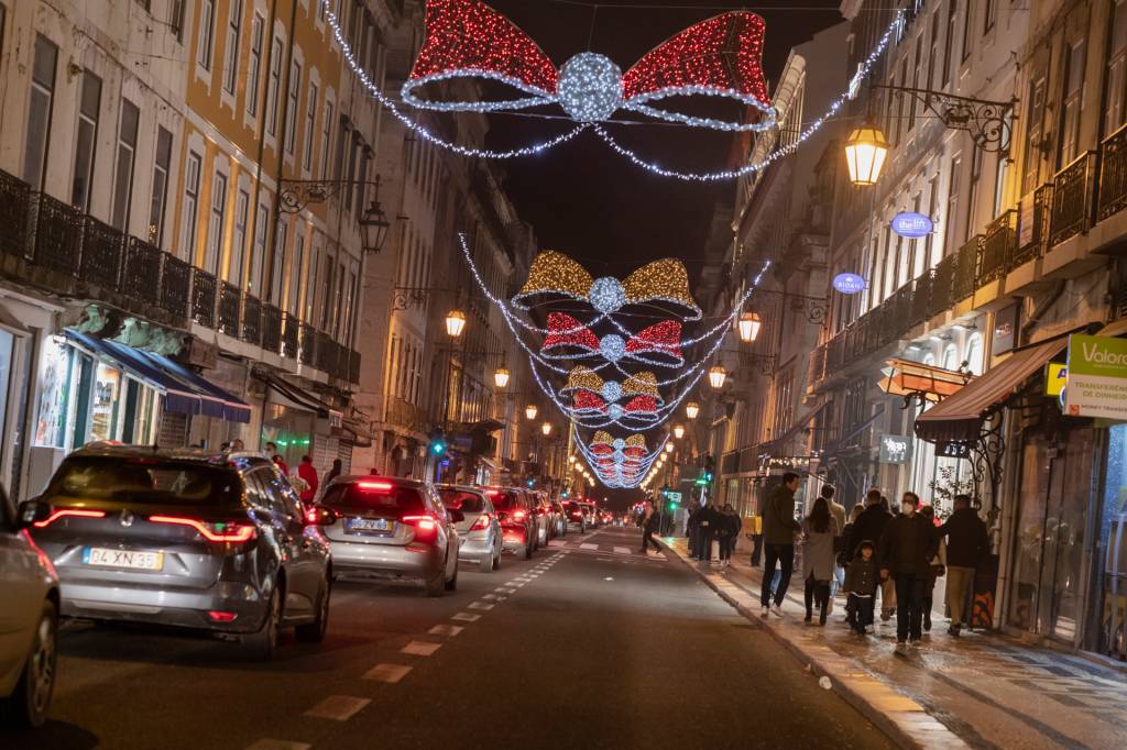 Rua com alguns carros passando, à noite, iluminada com grandes laços de luzes vermelhas e azuis para o Natal