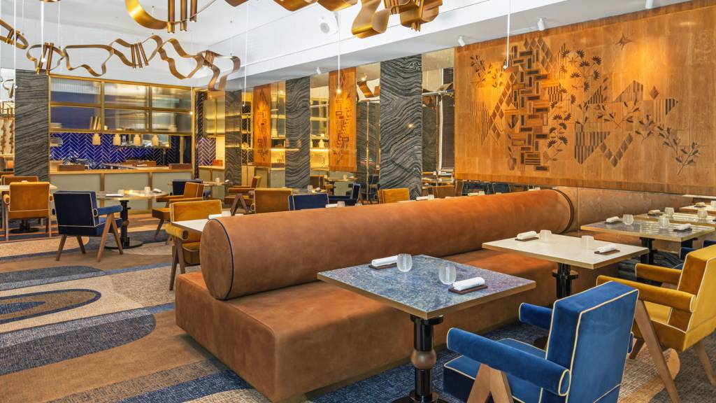 Salão do restaurante Cura onde se vê um sofá marrom com encosto cilíndrico ao centro, mesas de mármore e detalhes dourados na decoração