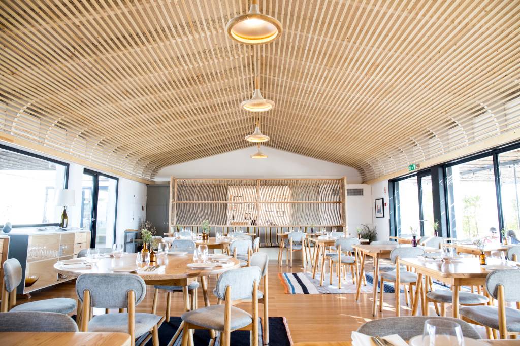 Salão de restaurante com teto em finas vigas de madeira, mesas e cadeiras em madeira clara com detalhes cinza e janelas de vidro que deixam entrar luz natural