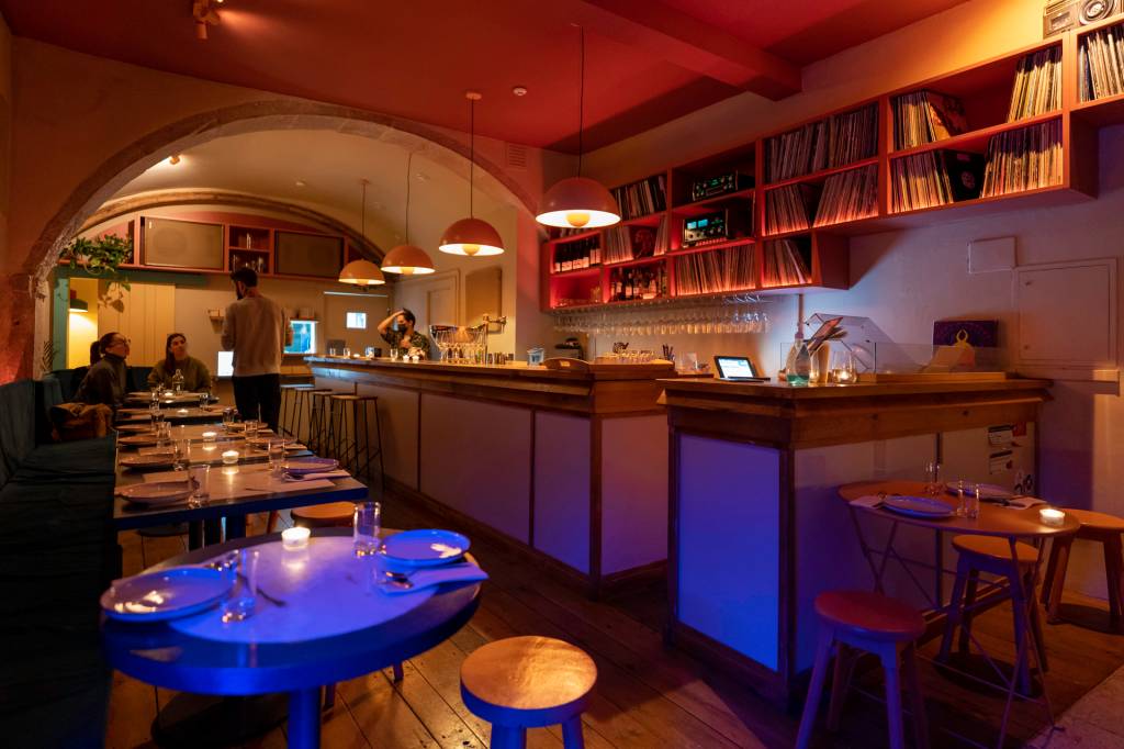 Bar estreito com iluminação em tons de azul, pequenas mesas com velas e um balcão sob quatro luminárias redondas