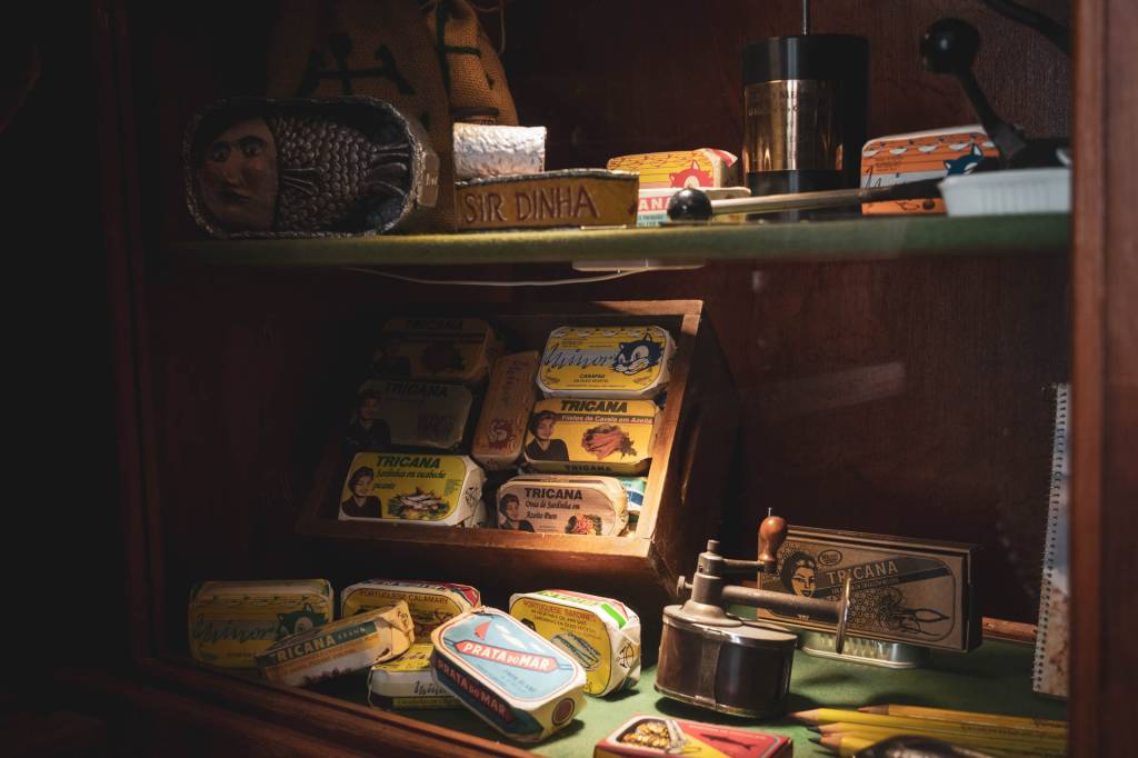 Uma vitrine em madeira com objetos antigos: latas de sardinhas e abridores