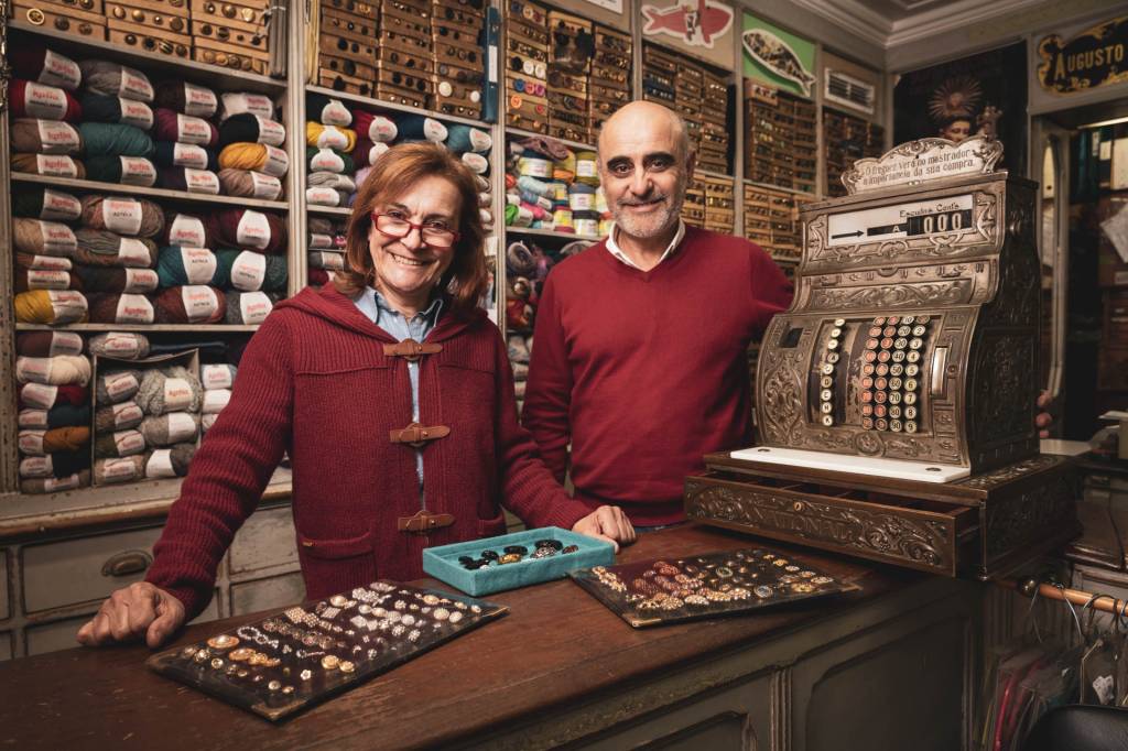 Um homem e uma mulher de blusas vermelhas posam para a fotografia num balcão de madeira de uma loja entre lãs e botões, ao lado de uma máquina antiga de calcular