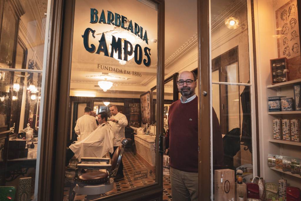 Um senhor careca, de barba, óculos e malha vermelha, abre uma grande porta de vidro onde está escrito Barbearia Campos