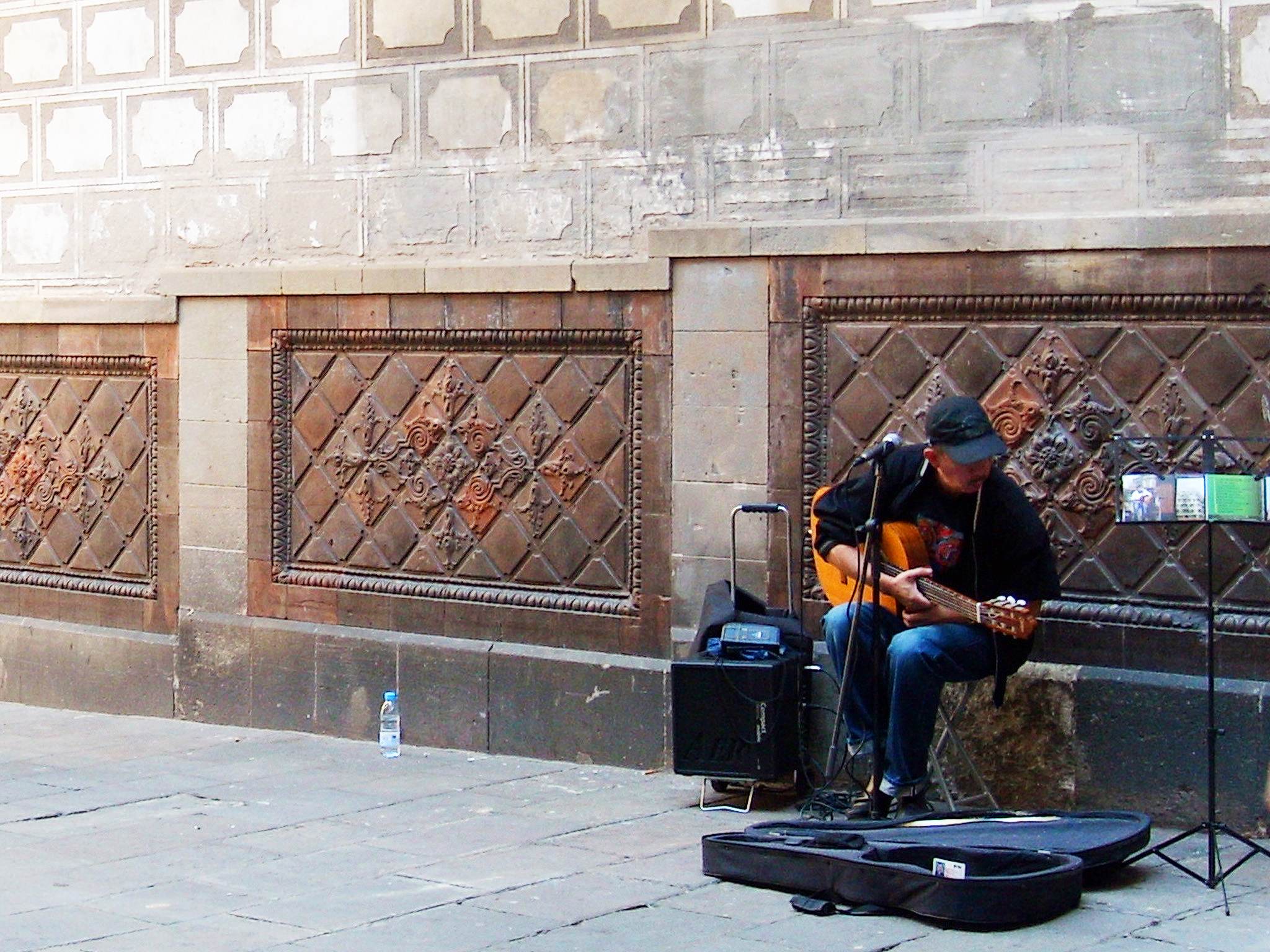 Homem tocando violão cuja caixa está no chão, aos seus pés. Ao fundo parede com arquitetura gótica.
