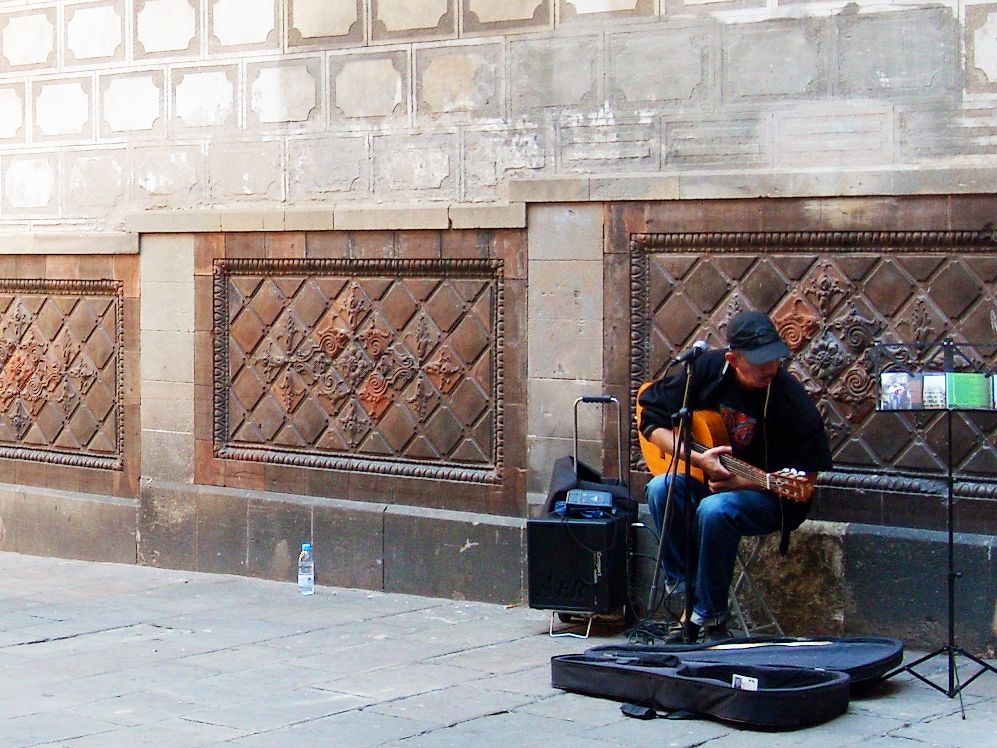 Barcelona Homem tocando violão cuja caixa está no chão, aos seus pés. Ao fundo parede com arquitetura gótica.