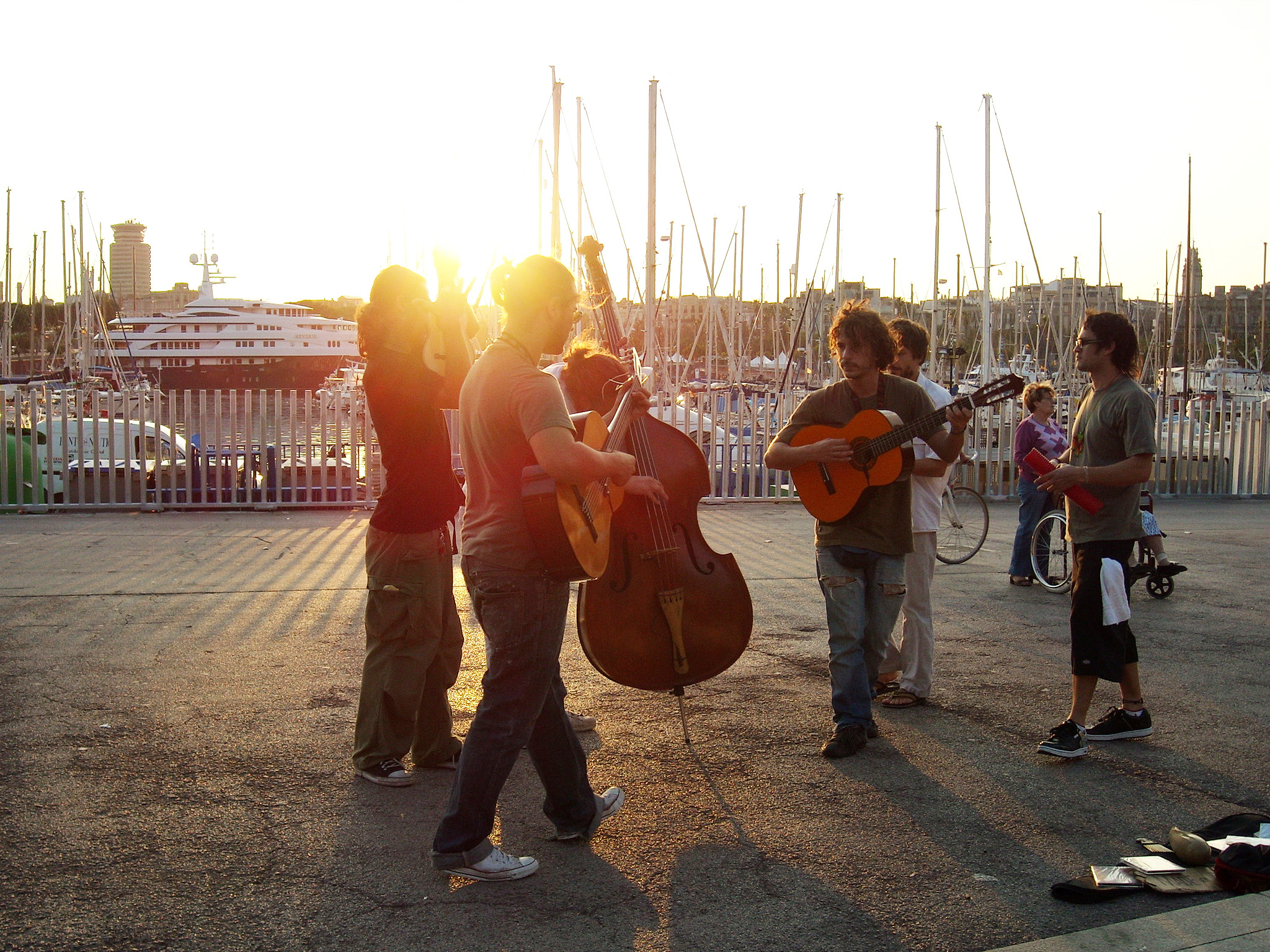 Músicos tocam diversos instrumentos no contra luz do pôr do sol de um porto com barcos ancorados ao fundo
