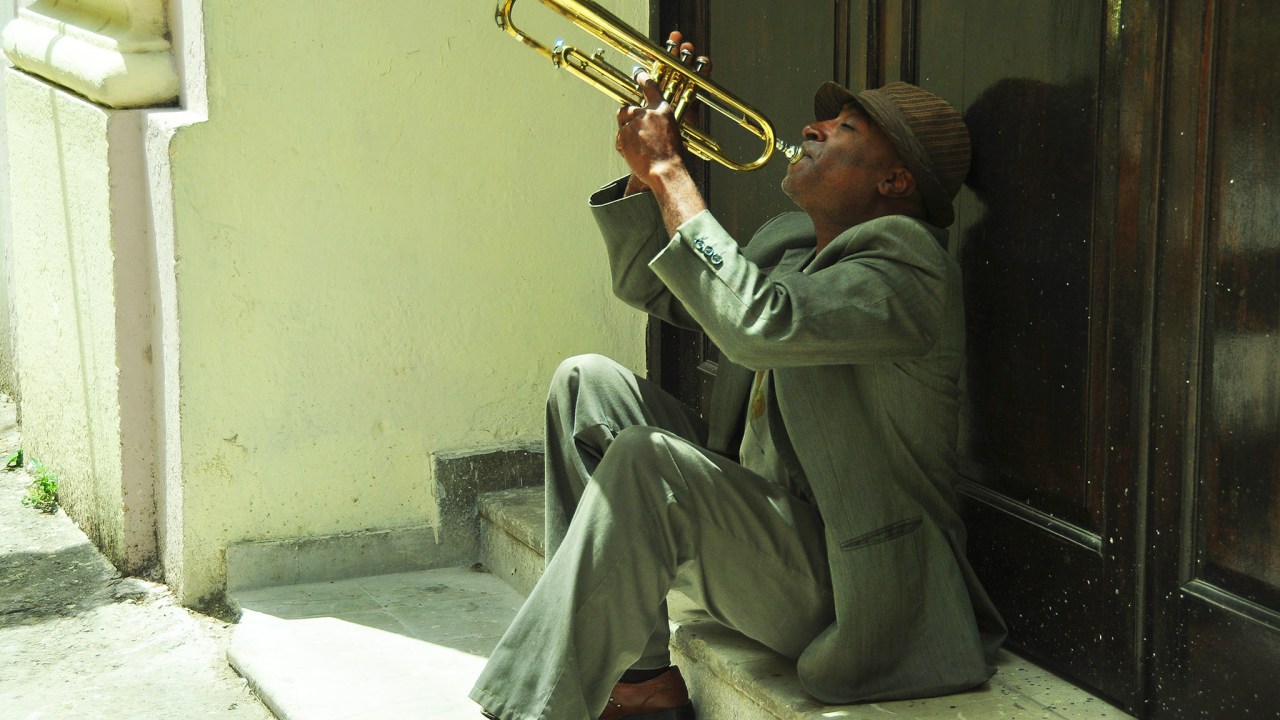 Homem preto sentado em degraus brancos e vestindo elegantemente um terno cinza e chapéu marrom, toca trompete com emoção.