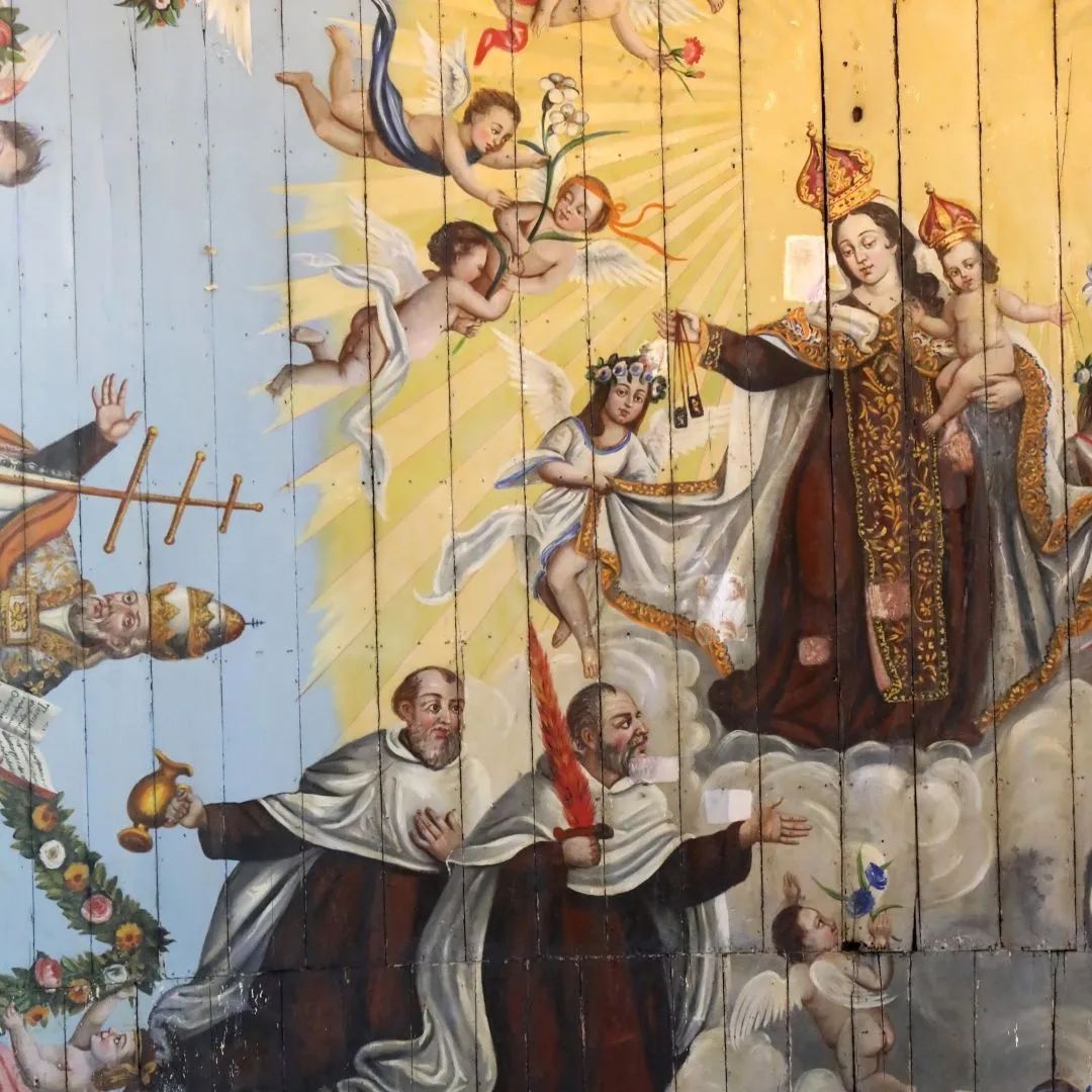 Na foto vemos uma pintura sacra feita sobre uma superfície de madeira. Vemos Nossa Senhora do Carmo carregando o menino Jesus e entregando o escapulário aos carmelitas.