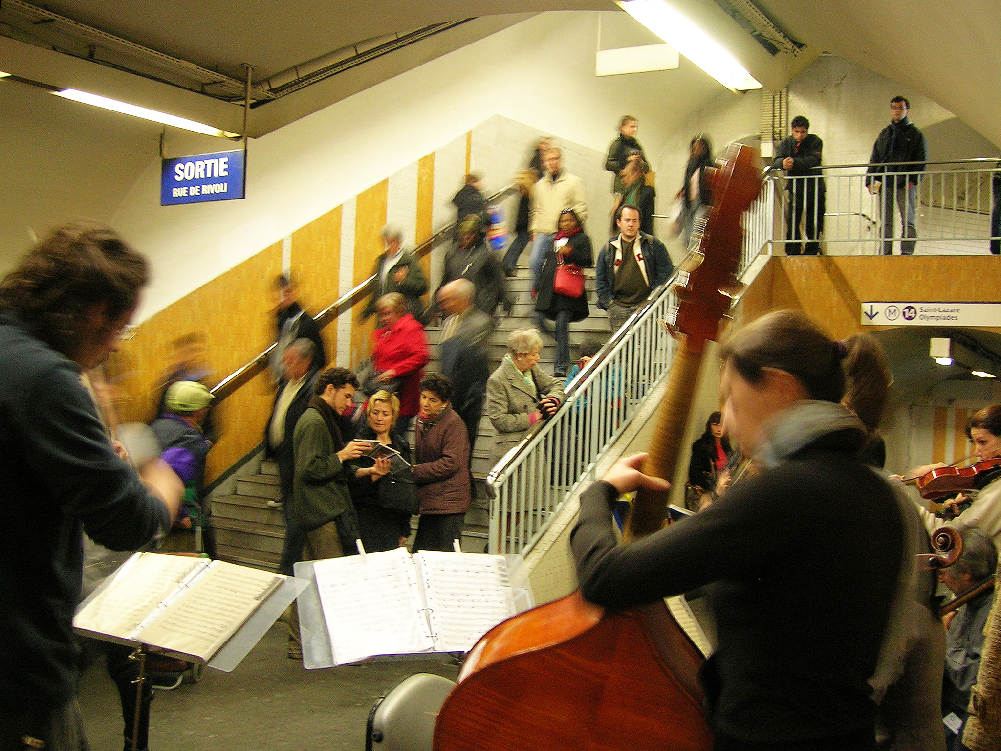 Paris Em primeiro plano um músico de costas toca um violoncelo. Ao fundo uma mulher toca violino. Pessoas descem a escada e três pessoas conversam paradas