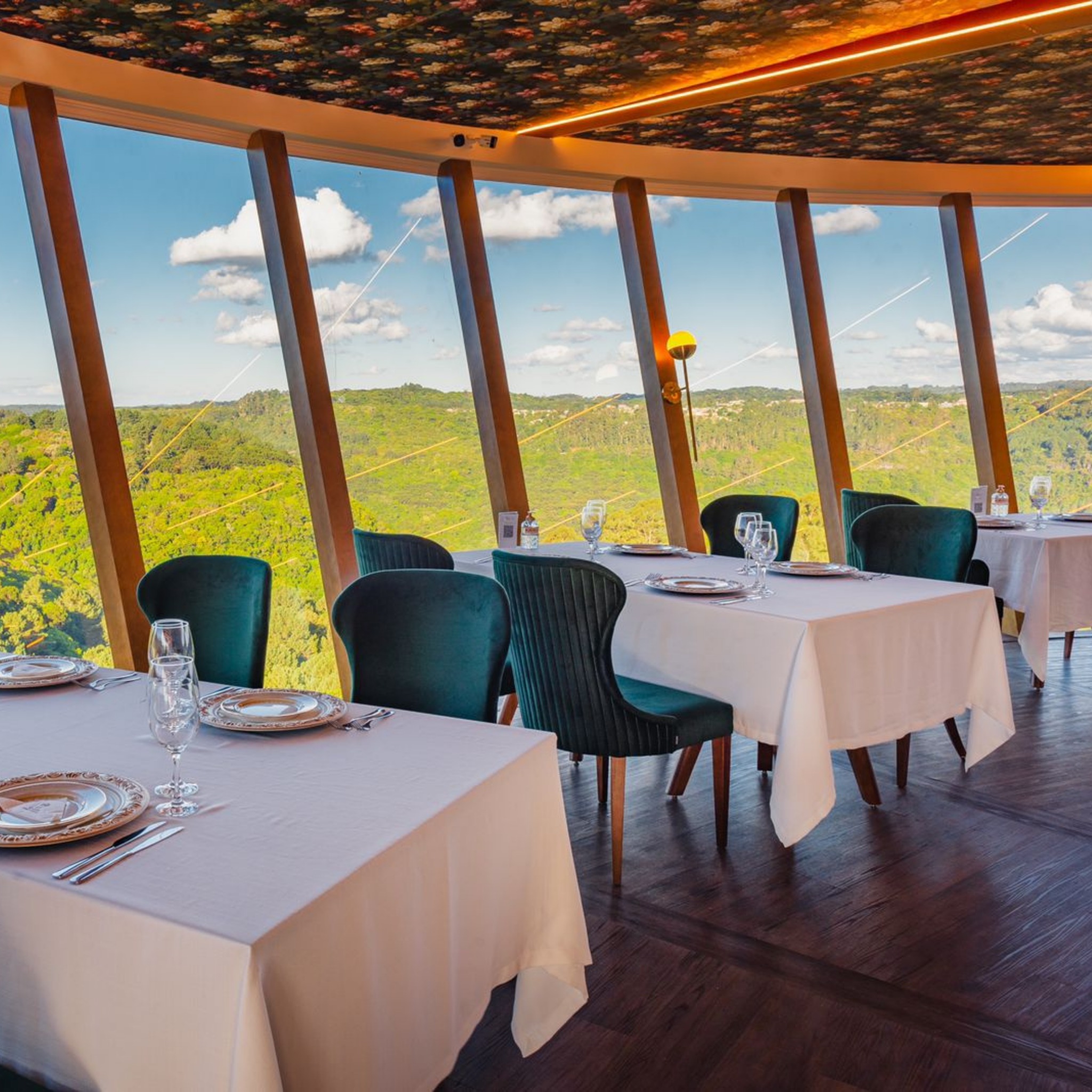Na imagem vemos o interior de um restaurante. Há várias mesas com toalhas brancas e cadeiras pretas. As paredes do restaurante são feitas de janelas, assim, vemos um vale verde que est[a ao lado do restaurante.