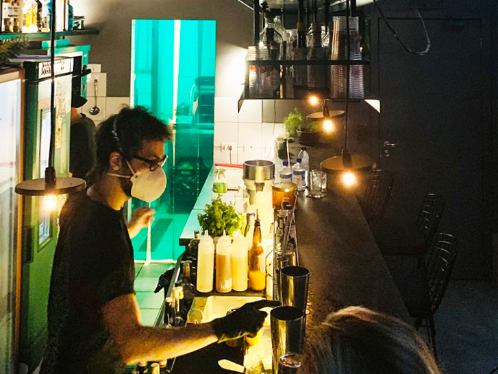Bartender usando máscara respiratória prepara bebidas no balcão de bar iluminado em contraste com o ambiente escuro