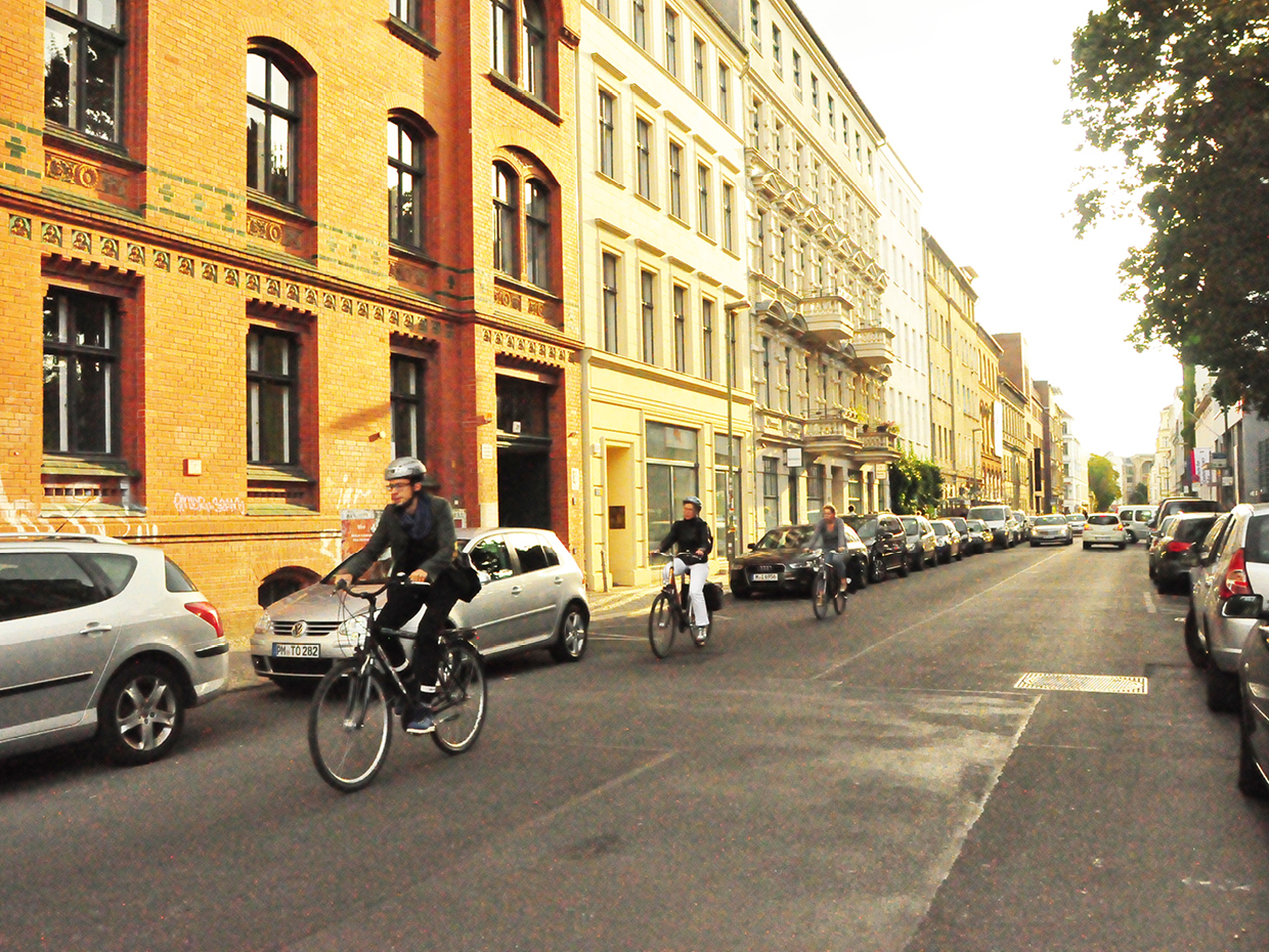 Rua do bairro de Scheunenviertel em Berlim com pessoas pedalando, carros estacionados e arquitetura da Berlim oriental.