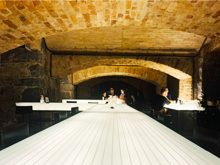 Em primeiro plano, como ponto de fuga: mesa de vidro iluminada. Pessoas ao fundo da imagem e arcos de tijolos no teto sobre as mesas.