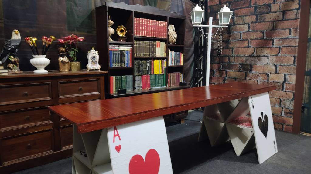 Na imagem vemos uma sala onde a mesa foi construída sobre gigantes cartas de baralho e as estantes estão cheias de livros antigos.