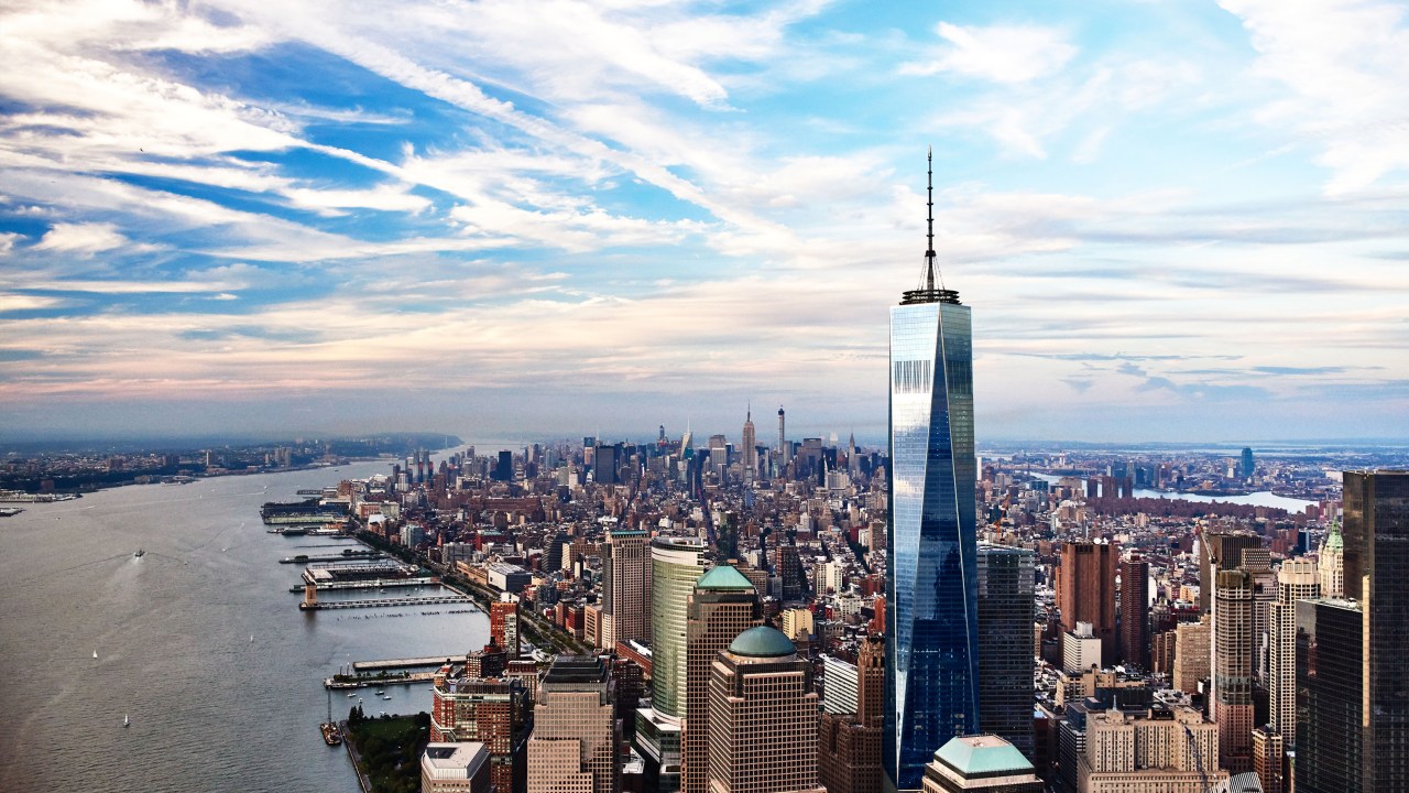 A figura é uma foto aérea de Nova York. Em foto, vemos o One World, um enorme prédio espelhado de azul, com uma torre alta.