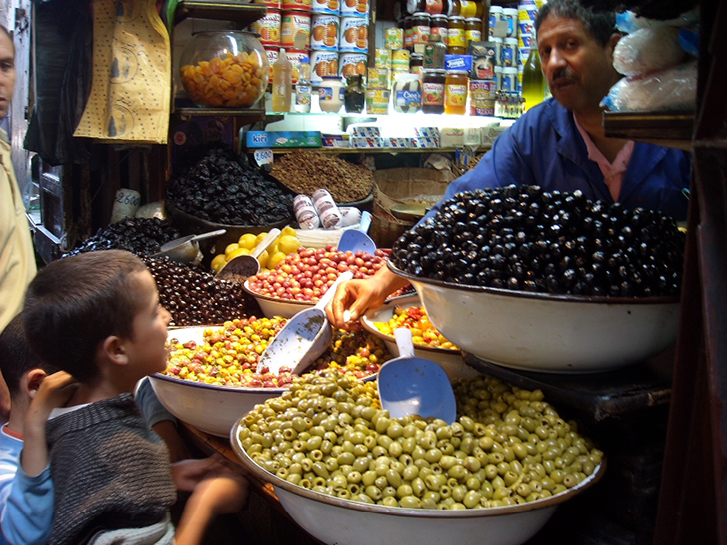 Dois meninos, um sorridente, compram azeitonas numa banca em Fez. O vendedor olha desconfiado direto para a câmera da fotógrafa