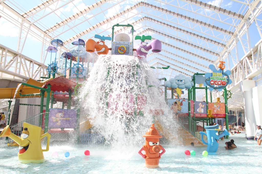 A imagem mostra uma área de piscinas infantis. É possível ver uma grande instalação de brinquedos em formato de torneiras, hidrantes e guarda-chuvas, além de um grande balde que despeja água sobre a rasa piscina.