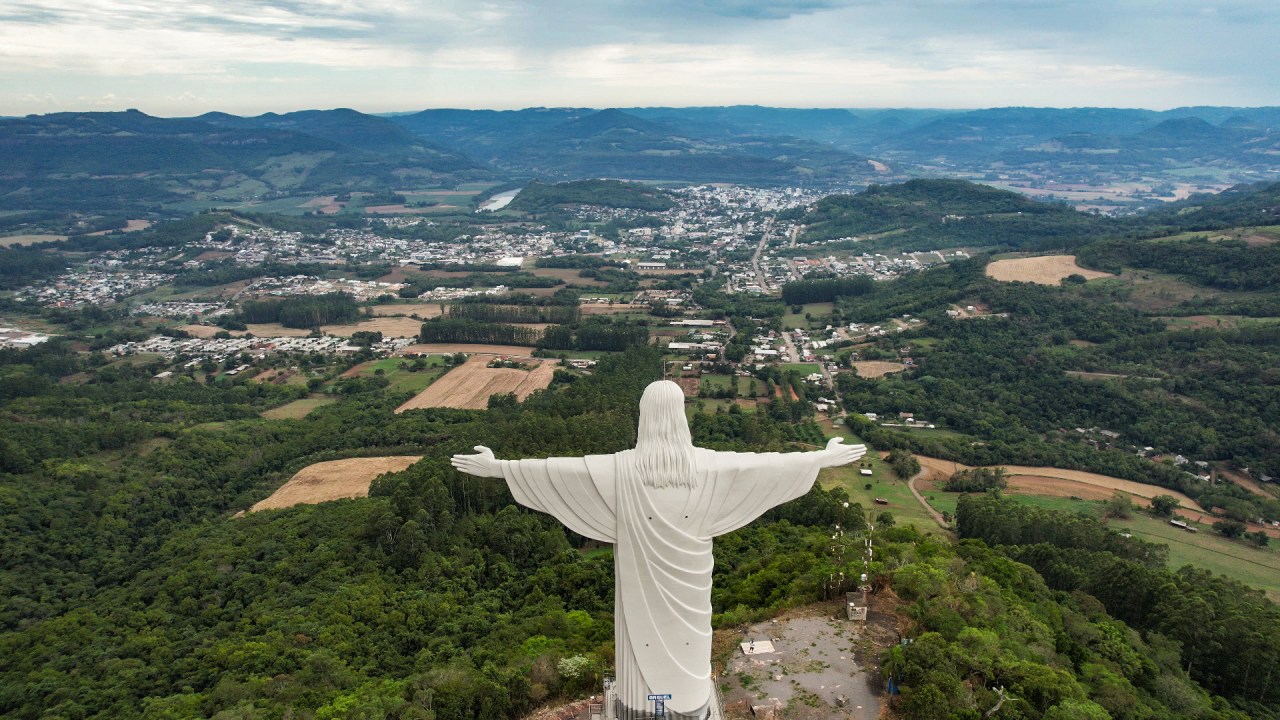 A imagem mostra uma estátua cinza claro do cristo de costas, com os braços abertos. Ao fundo é possível ver um vale e uma cidade.