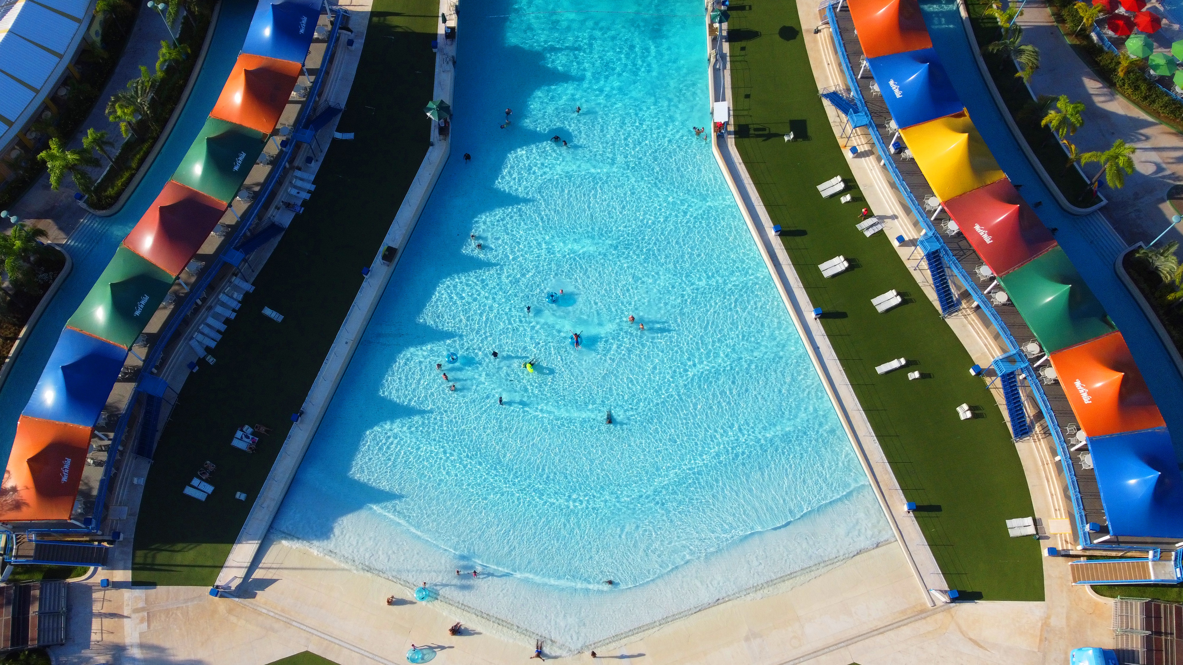 A imagem mostra um,a piscina de ondas vista de cima. Ao redor da atração, é possível ver tendas de diversas cores.
