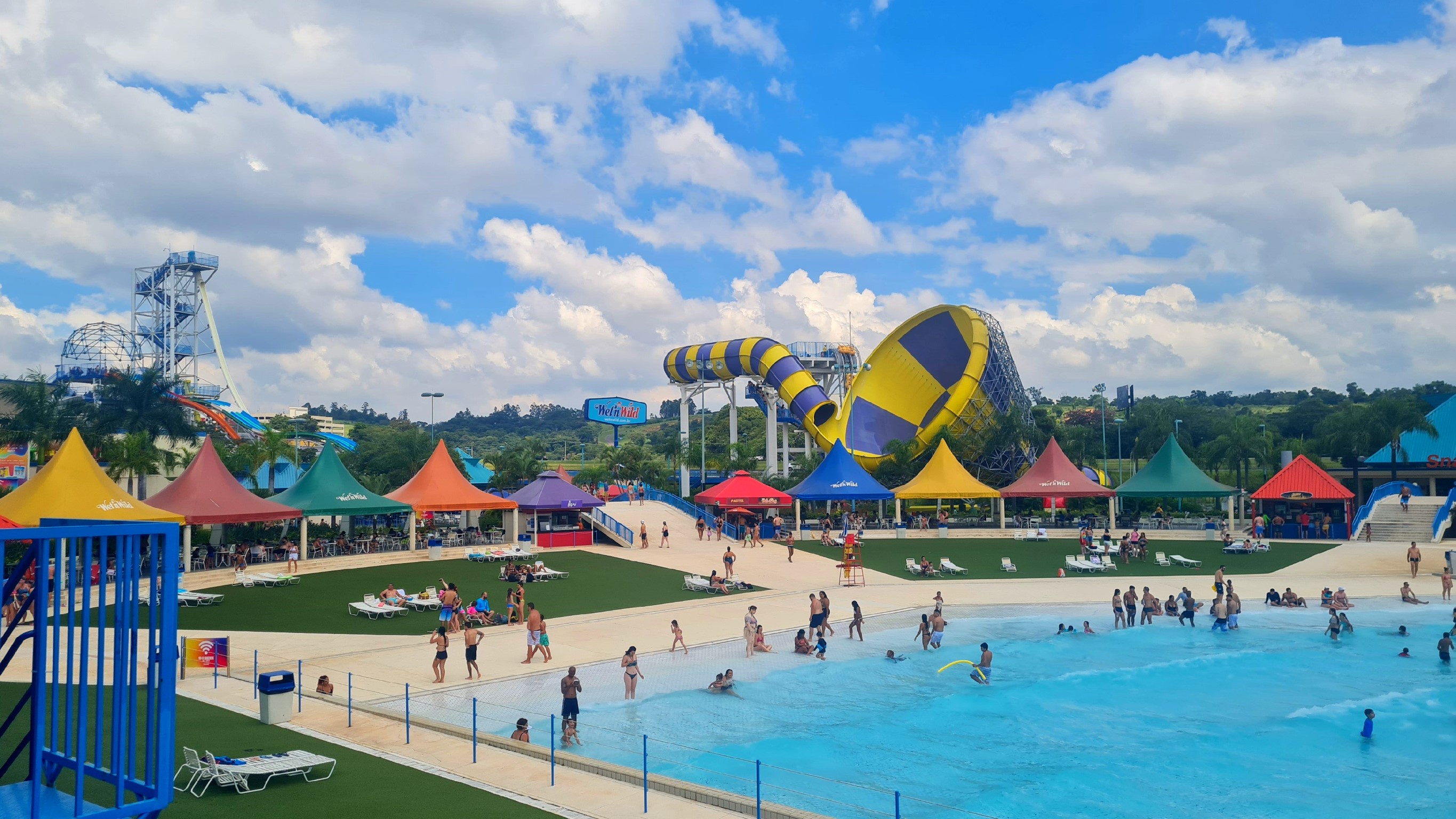 Na fotografia é possível observar uma piscina de ondas cercada por tendas coloridas. Ao fundo, vê-se um enorme brinquedo que se parece com um funil amarelo e roxo.