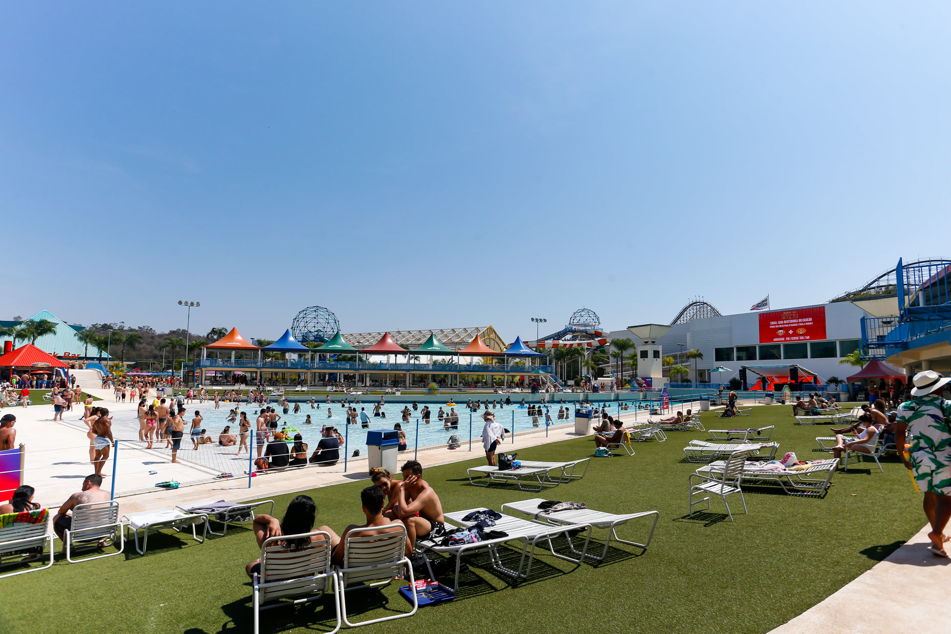 A imagem mostra várias pessoas em espreguissadeiras ao lado de uma piscina de ondas.