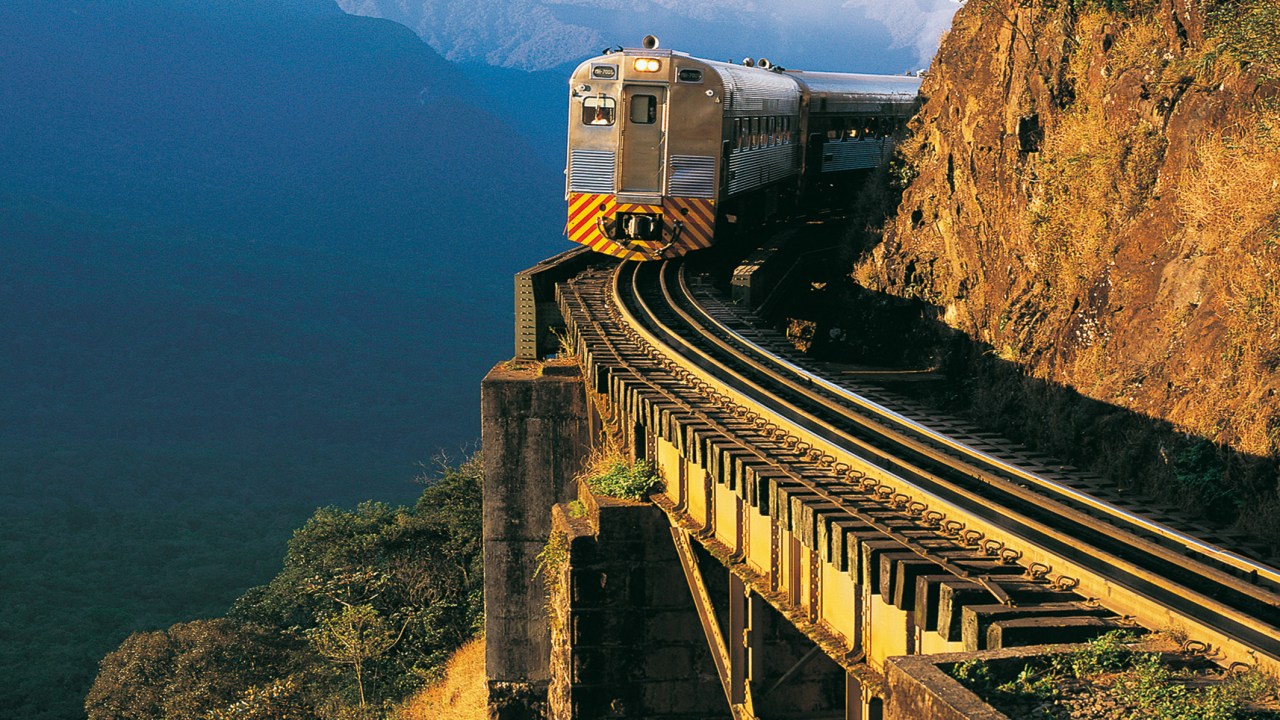 A imagem mostra um trem sobre trilhos curvados. A curva segue o alinhamento de uma montanha ao lado direito, enquanto o lado esquerdo possui um abismo. Ao fundo, a sombra de outras montanhas é azul escuro