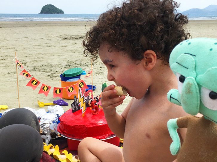 Um menino de 5 anos em primeiro plano, comendo. O cenário: uma festa na praia, com bolo, decoração e doces. Ao fundo a praia de Ubatumirim