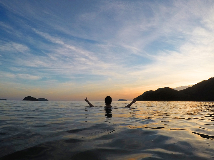 A woman in a meditation sign immersed in the sea to her neck with her arm raised.  In the background, pink sunset