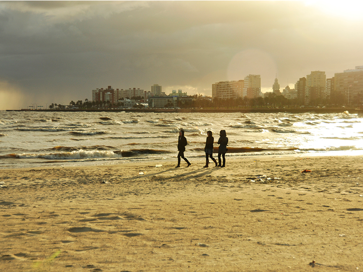 Tres pessoas andando na praia num contraluz de fim de tarde. Ao fundo o skyline da cidade de Montevideo