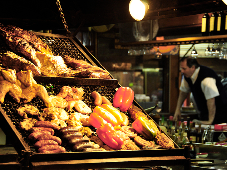 Grelha de parrilla com carnes e legumes sendo grelhados. Ao fundo um homem desfocado