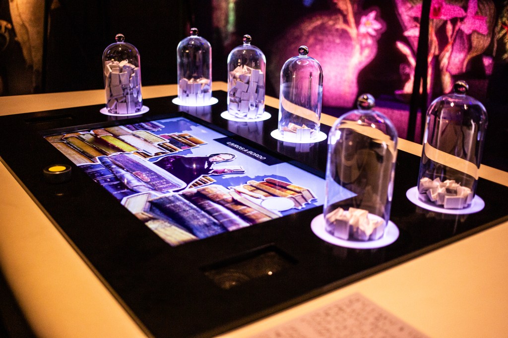 A imagem mostra uma mesa com uma tela e vidros cheios de papéis dobrados.