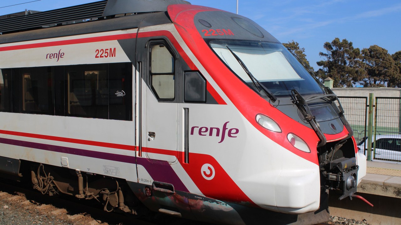 A imagem colorida mostra um trem moderno branco e vermelho, com o logo da empresa Renfe