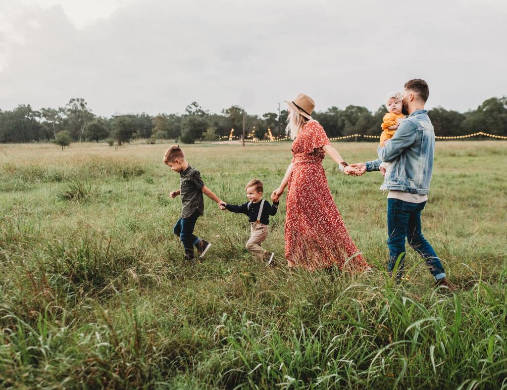 Uma família com a mãe de vestido vermelho e chapéu, o pai de jaqueta e calça jeans, um bebé e duas crianças caminha por um campo de plantas verdes
