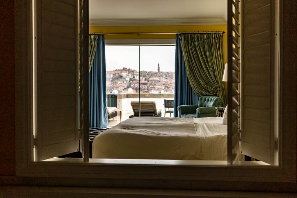 Quarto de hotel com uma janela de madeira que se abre para a cama e, no fundo, a varanda com vista da cidade do Porto