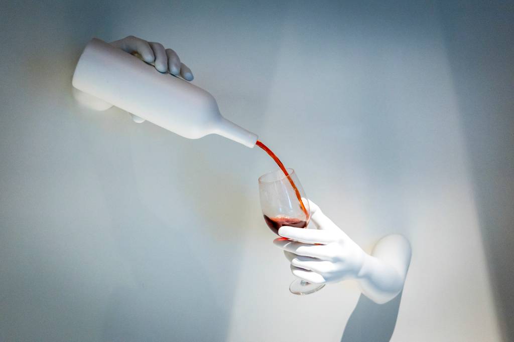 Escultura em parede branca mostra uma mão segurando uma garrafa, também brancas, servindo vinho tinto a uma taça segurada por outra mão